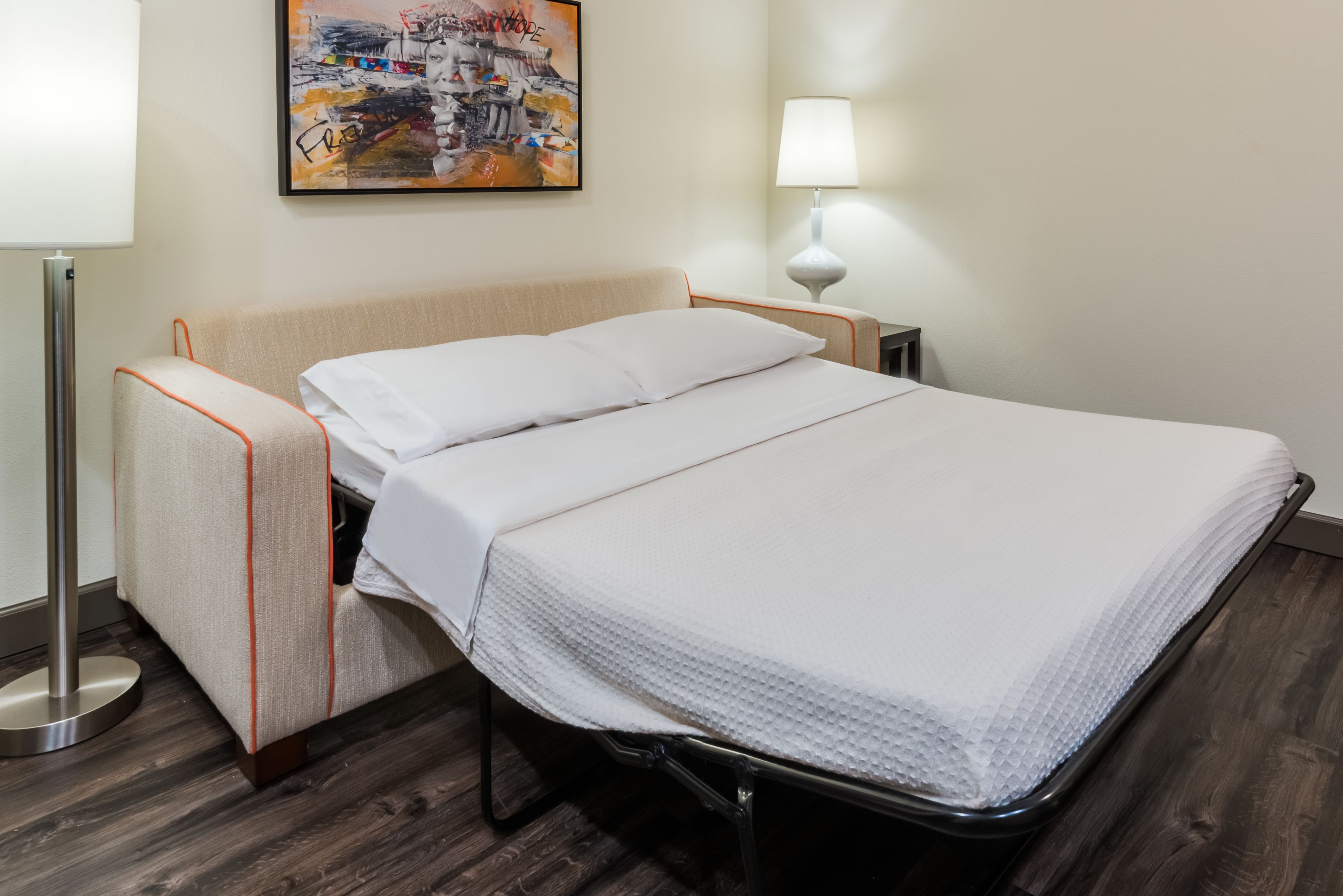 A neatly made bed with white linens, a bedside lamp, and a framed artwork on the wall.
