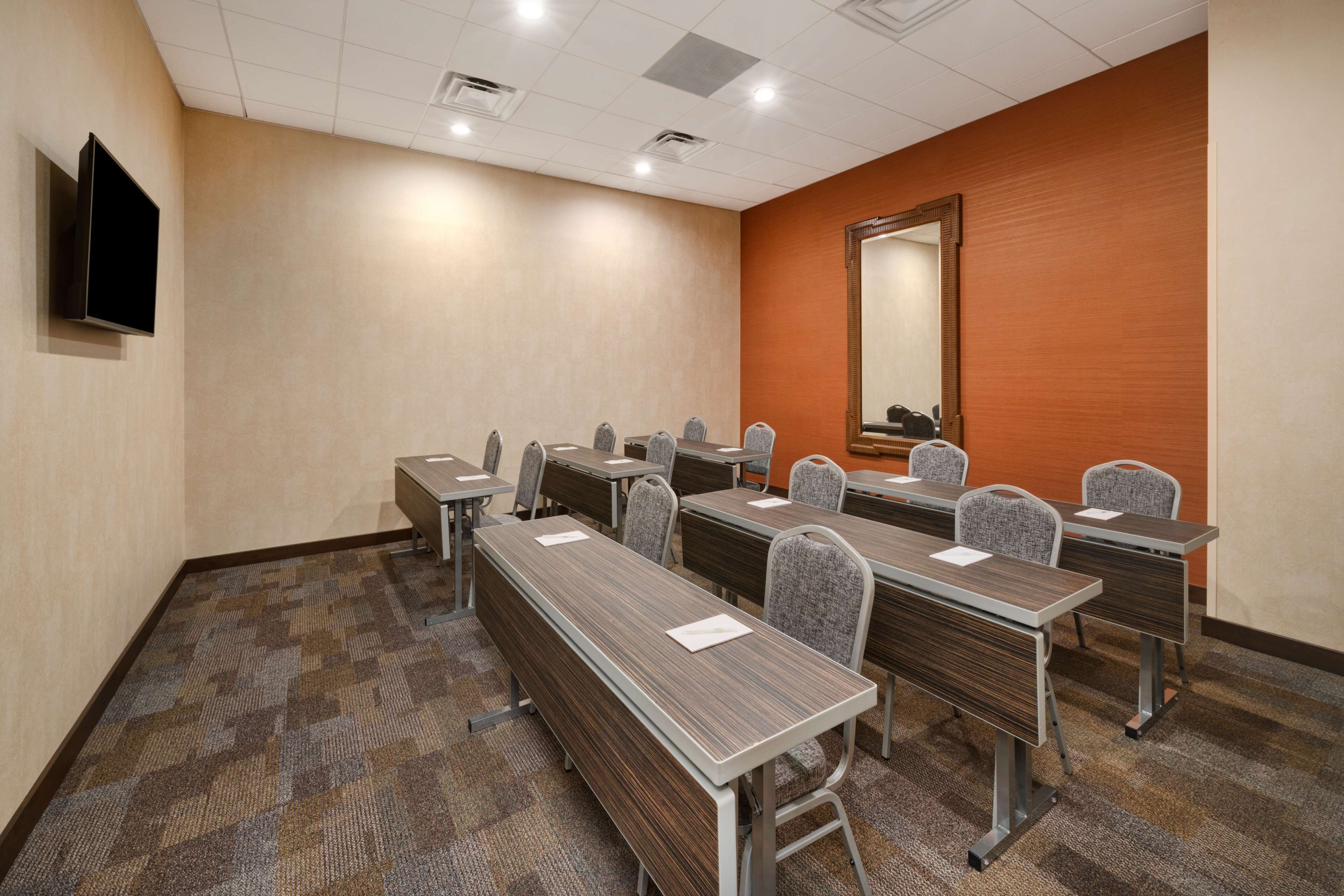 A conference room with multiple tables and chairs, a flat-screen TV mounted on the wall, and a large mirror on the orange wall.