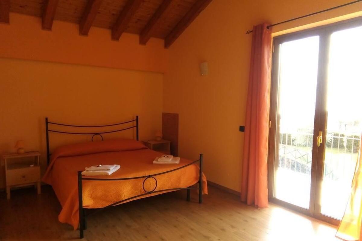 Un dormitorio con cama, suelo de madera, una mesita de noche, una lámpara y un ventanal corredero al exterior.