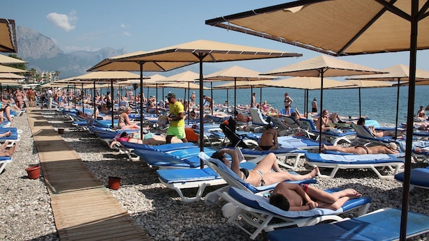 A beach with rows of sun loungers and umbrellas, a wooden walkway, and people sunbathing.