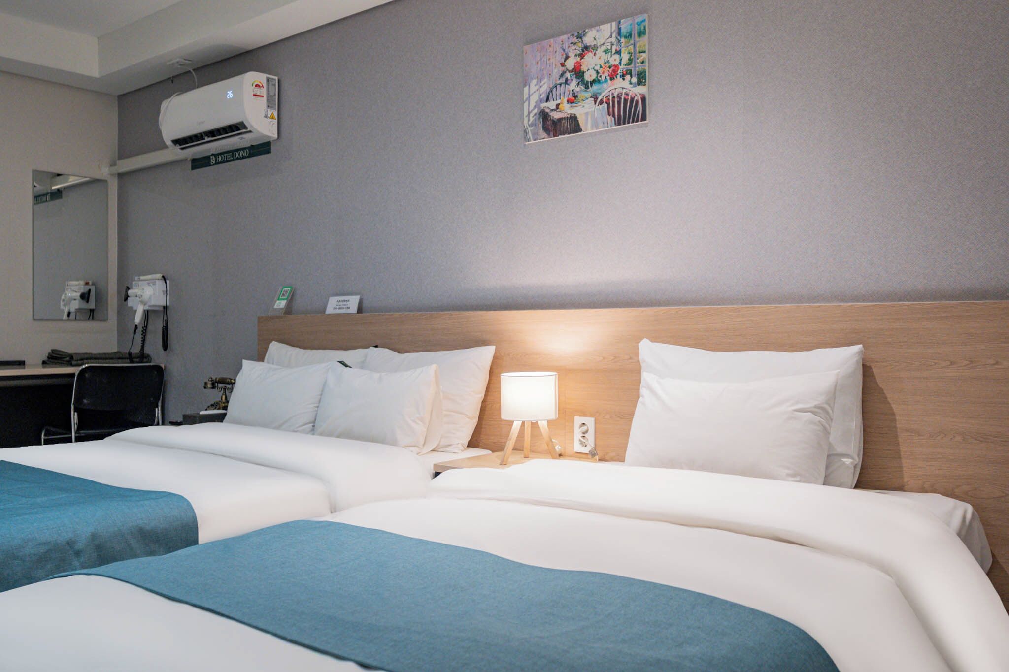 A hotel room with two beds, a desk with a chair, a mirror, an air conditioning unit, and a painting on the wall.