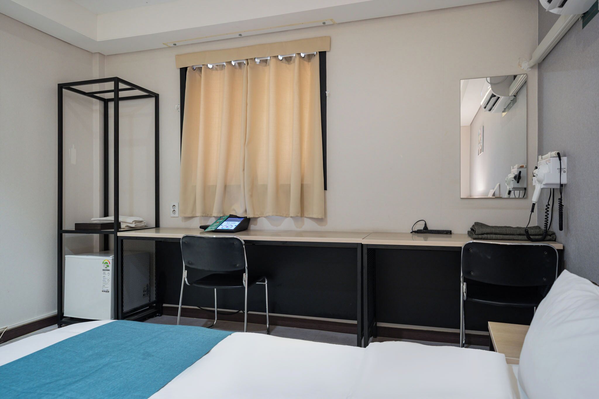 Une chambre d’h ôtel comprenant un lit, un bureau, deux chaises, un petit réfrigérateur, un miroir et une fenêtre avec des rideaux.