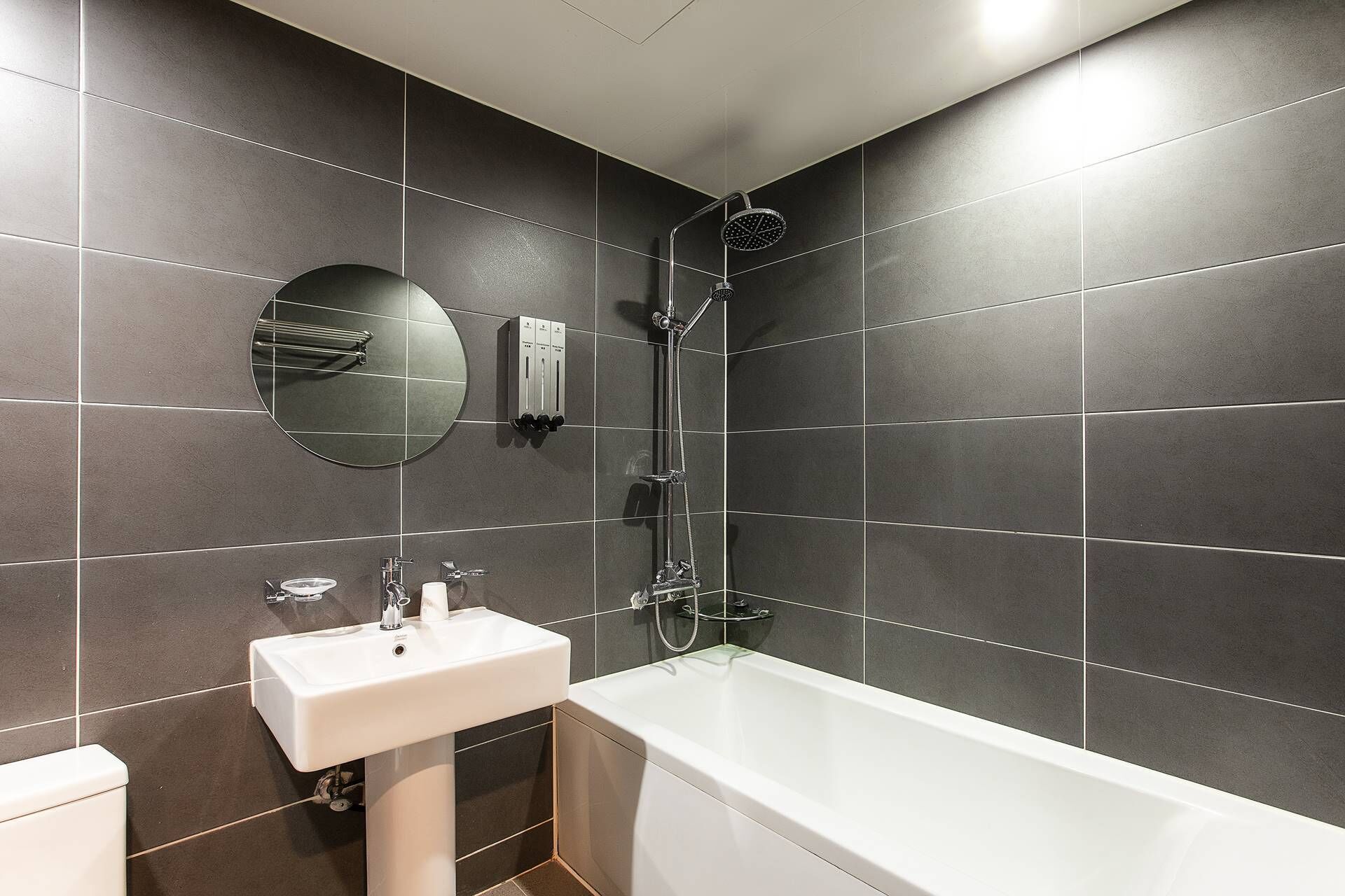 A modern bathroom with dark tiles, a white sink, a round mirror, and a walk-in shower.