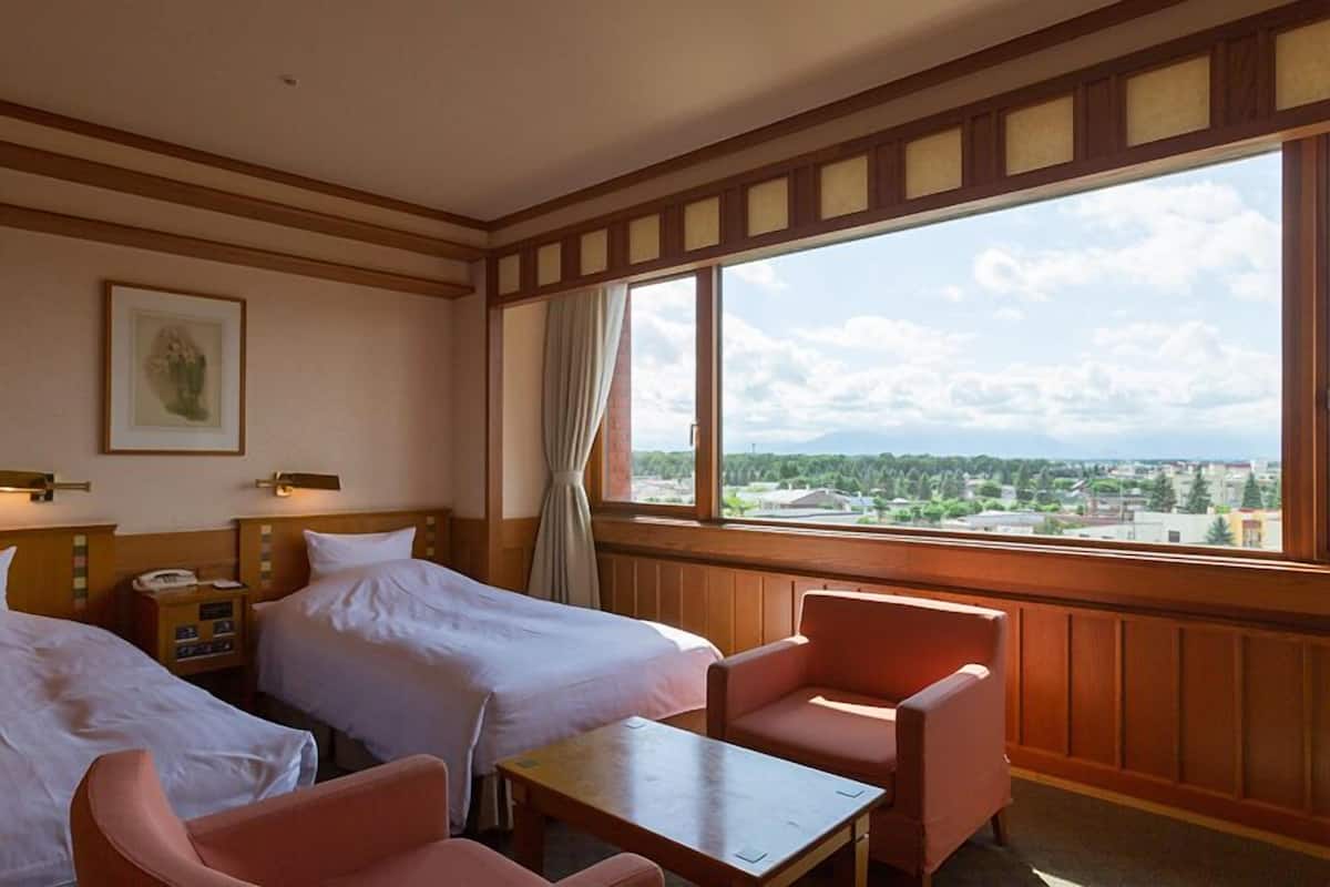 Ein Hotelzimmer mit zwei Betten, einem kleinen Tisch, einem Stuhl und einem großen Fenster mit Blick auf die Stadt.