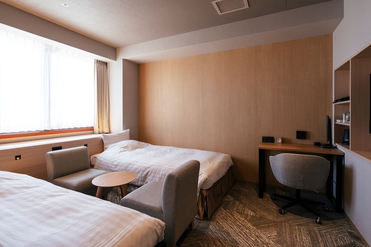 Ein Hotelzimmer mit zwei Betten, einem Schreibtisch, einem Stuhl, einem kleinen runden Tisch und einem großen Fenster mit Vorhängen.