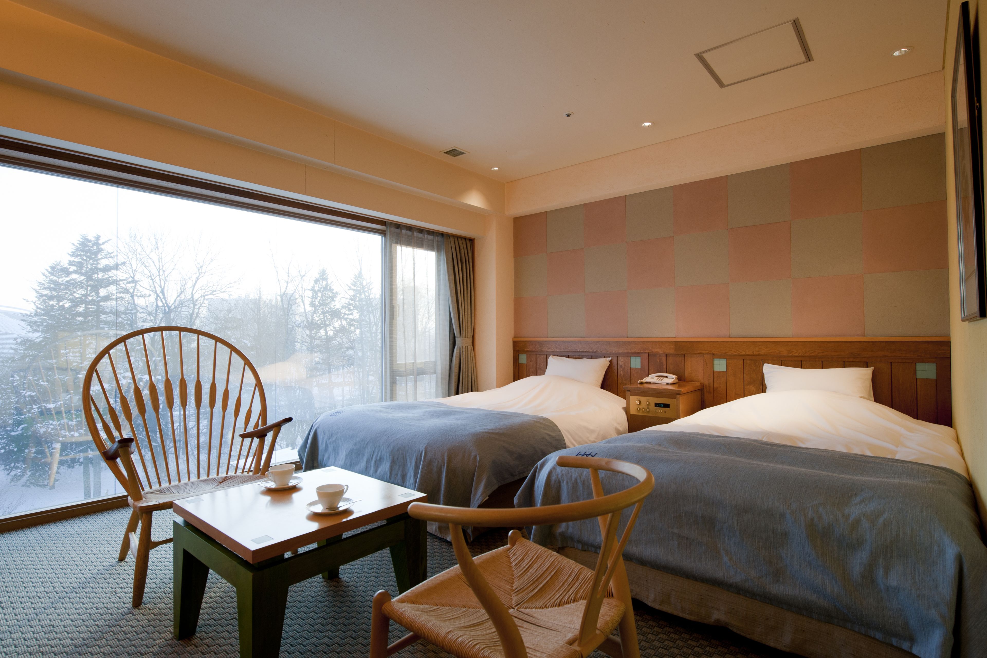 A hotel room with two beds, a wooden chair, a table, and a view of trees through the window.