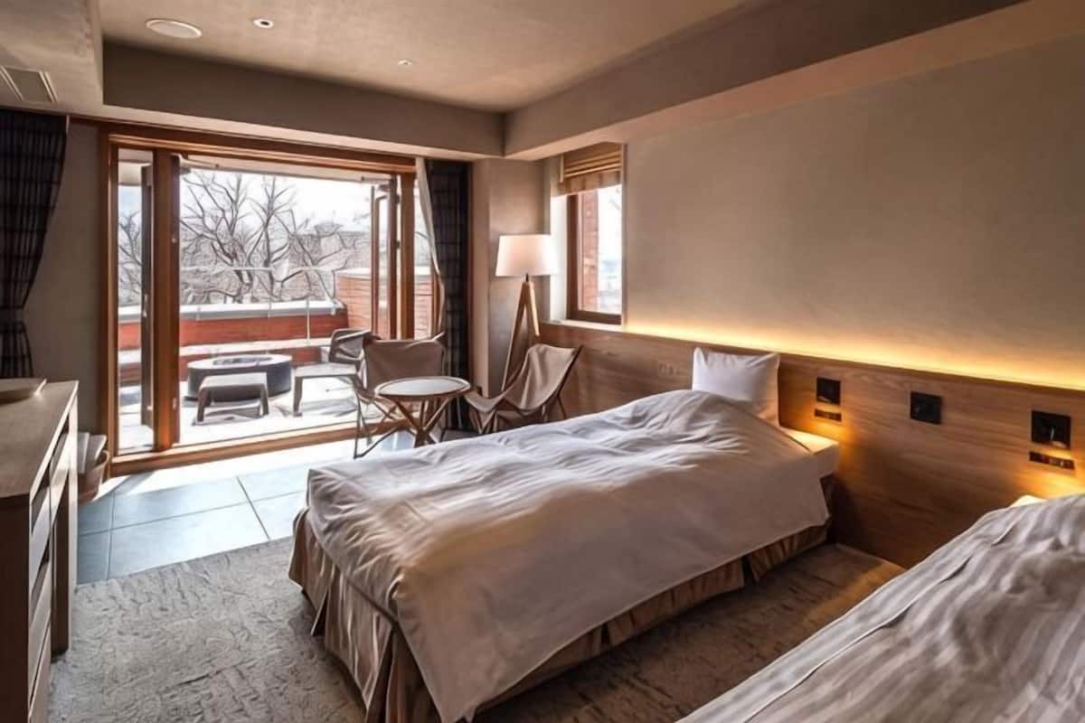 A hotel room with two beds, a desk, a chair, a small table, and a view of the outdoors.