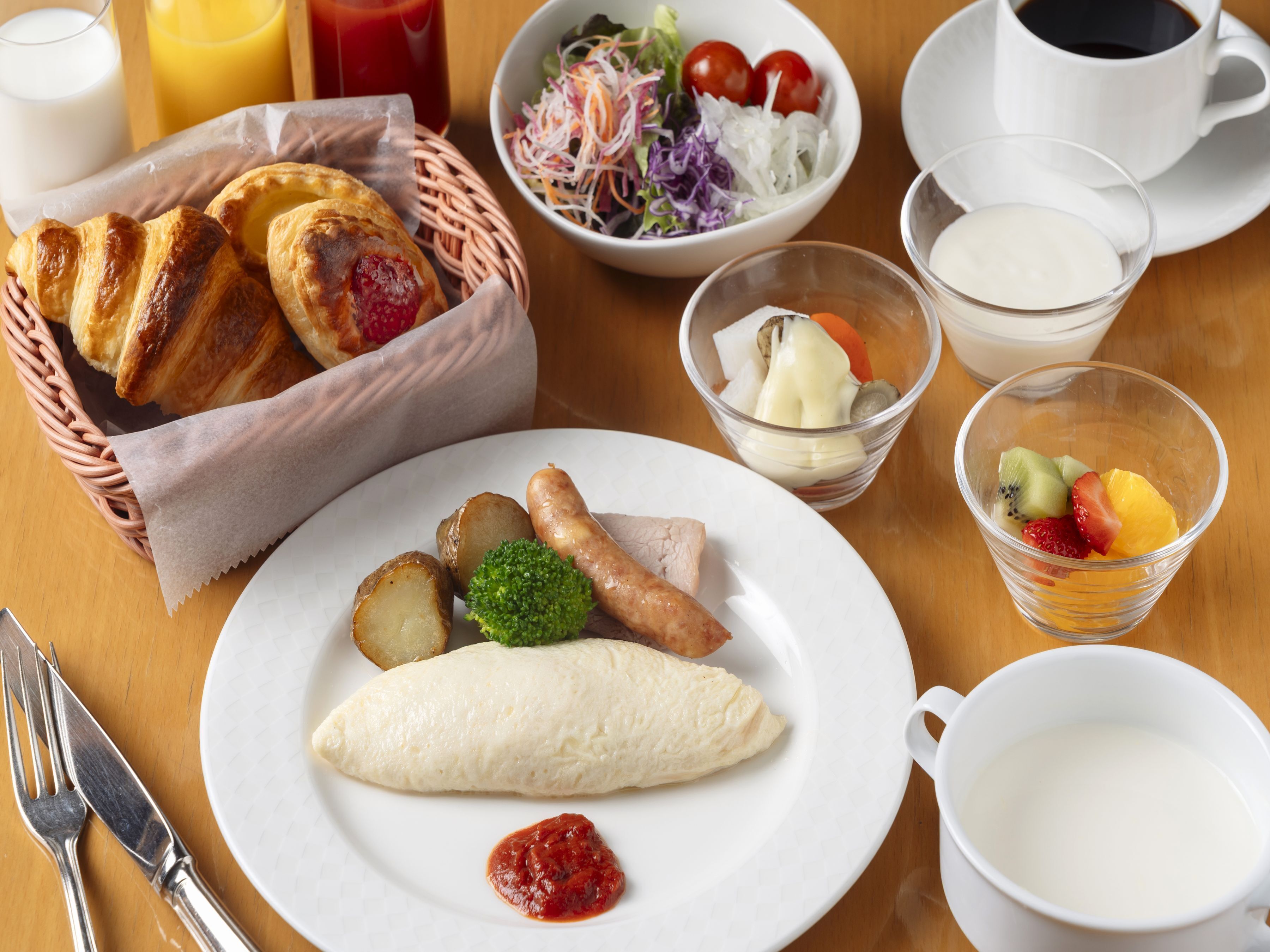 Une table dressée pour le petit-déjeuner avec des croissants, une assiette de saucisses, des pommes de terre et un œuf dur, ainsi qu’une salade de fruits, un bol de salade et une tasse de café.