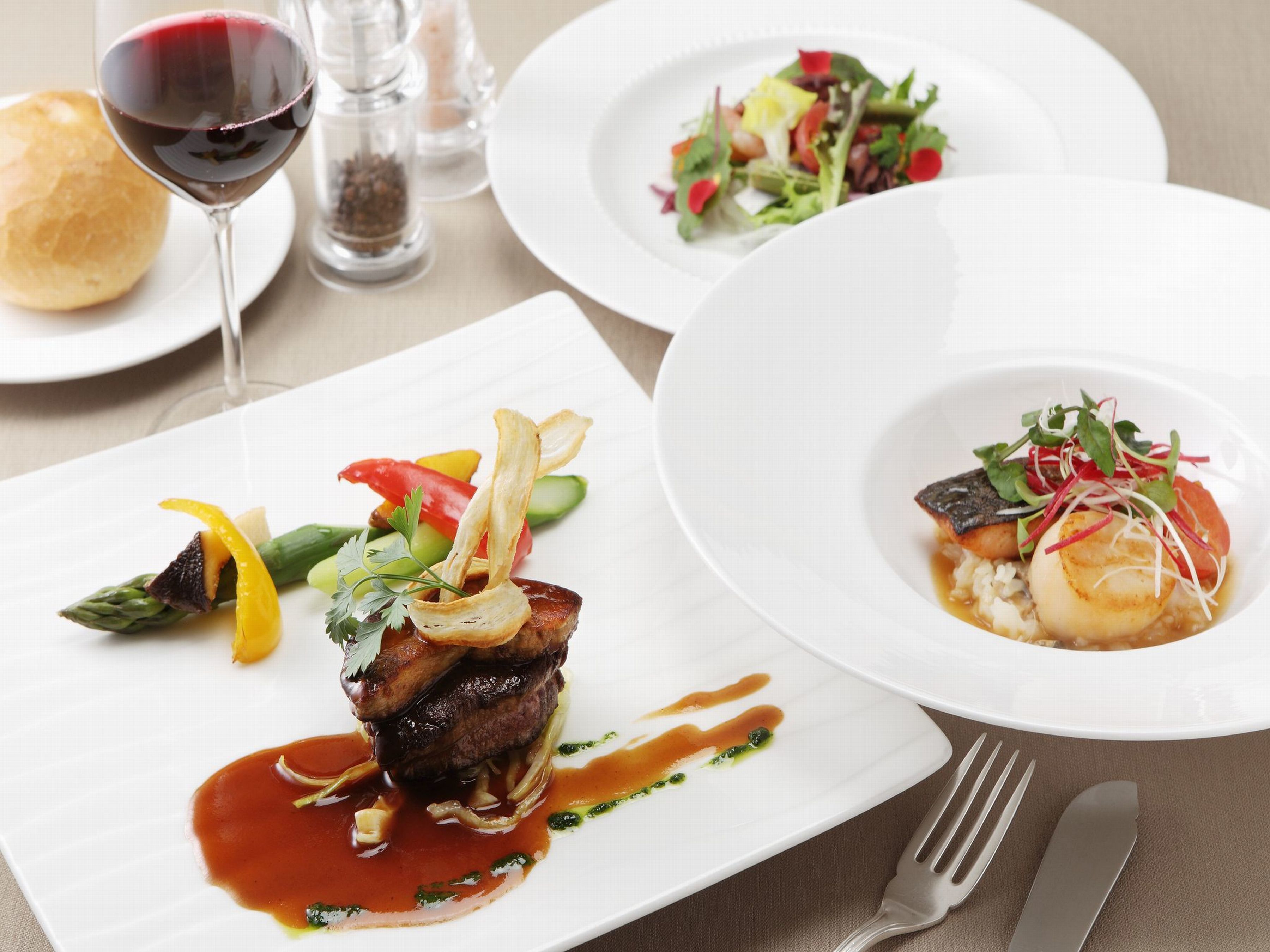 A dining table with three plates of food, a wine glass, and a salt and pepper shaker.