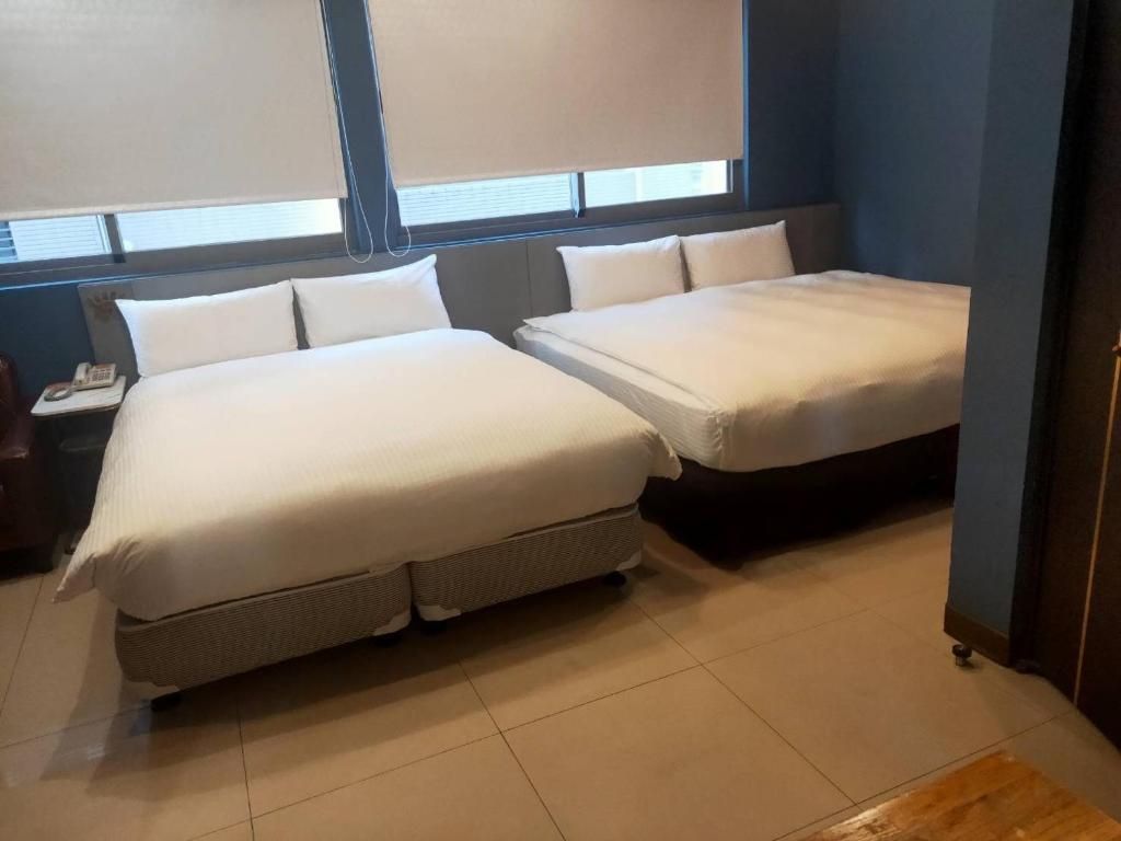 A hotel room with two beds, a nightstand with a phone, and a window with blinds.