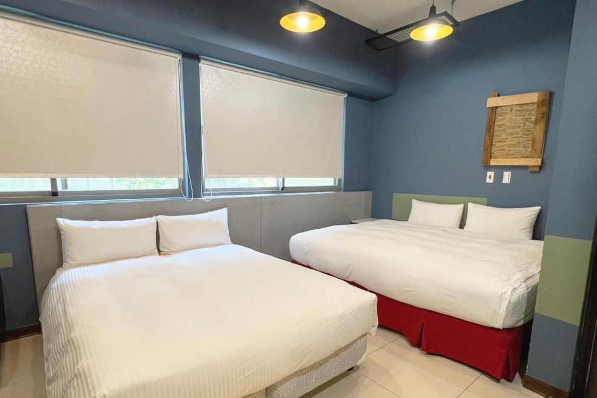 Habitación de hotel con dos camas, una ventana con persianas, una lámpara de pared y una placa de madera.