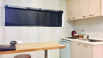Een compacte keuken met een houten tafel en een krukje, een kleine koelkast en lichtkleurige kastjes.