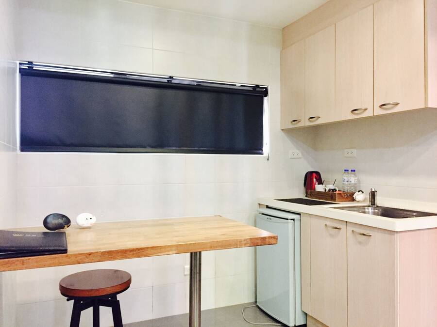 Een compacte keuken met een houten tafel en een krukje, een kleine koelkast en lichtkleurige kastjes.