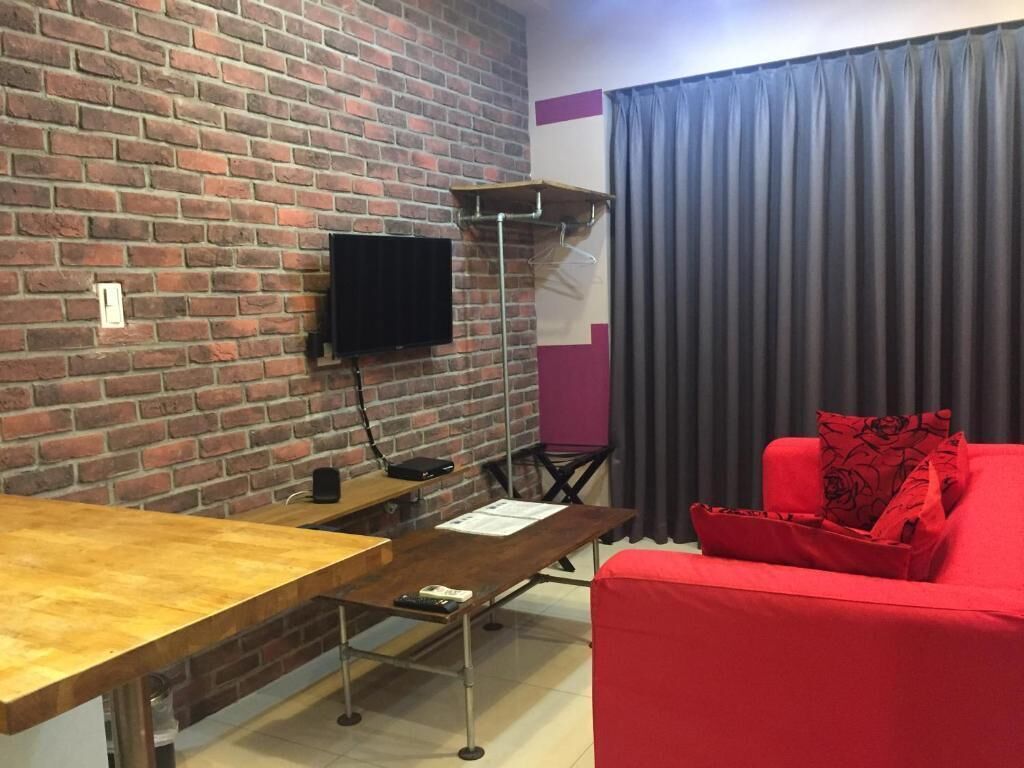 A room with a brick wall, a flat-screen TV mounted on it, a wooden table, a red sofa, and a grey curtain.