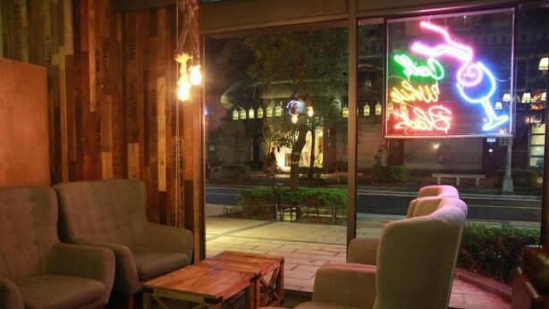 Un espacio interior acogedor con sillas acolchadas y una mesa de madera, un letrero de neón en el exterior y vistas de la calle a través de amplios ventanales.