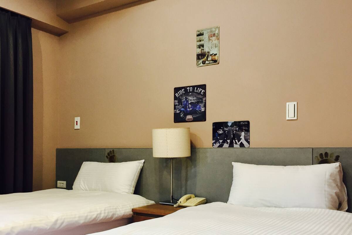 Une chambre d’hôtel avec deux lits, une lampe de chevet, un téléphone et des éléments de décoration murale, notamment une affiche intitulée « Ride to Life » et une photographie intitulée « Cross the Pond ».
