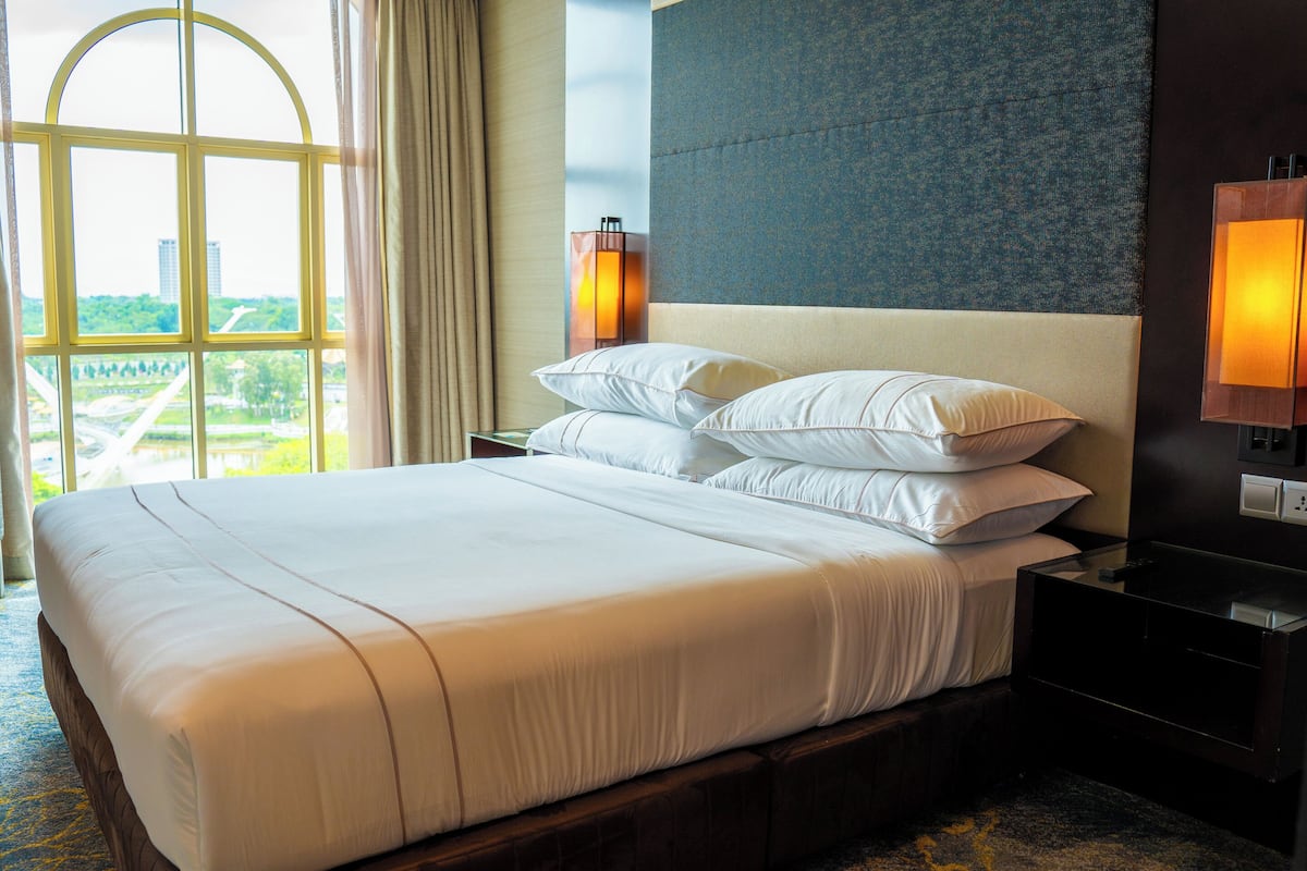 A hotel room with a large bed, two bedside lamps, a view of a cityscape, and a window with curtains.