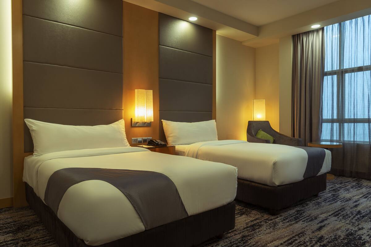 A hotel room with two beds, a large window with curtains, and a wall-mounted lamp.