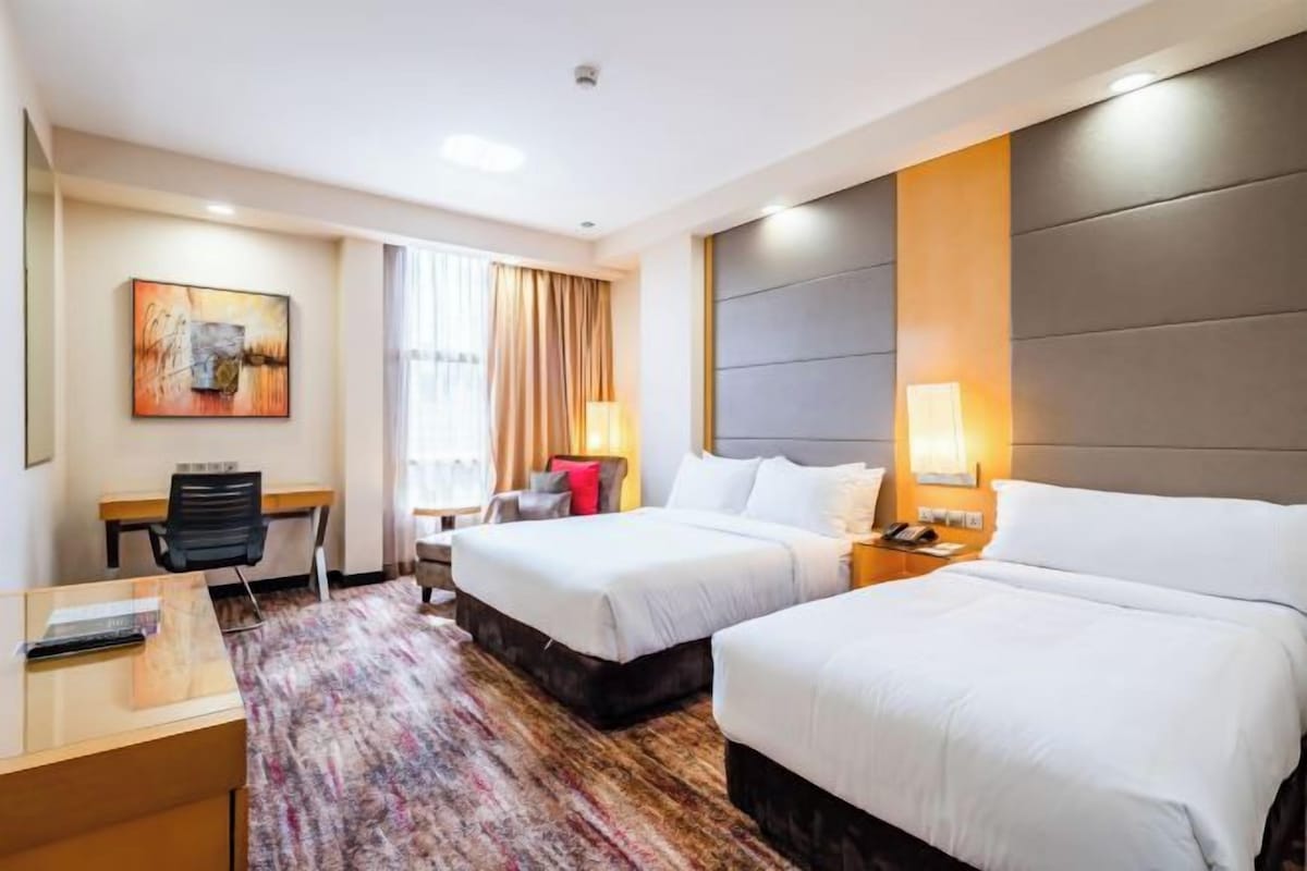 Habitación de hotel con dos camas, un escritorio, una silla, un cuadro grande y una ventana con cortinas.