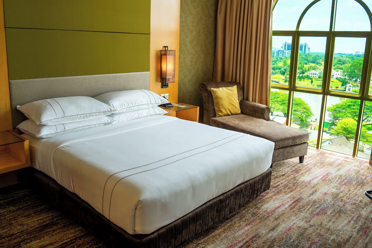 A hotel room with a large bed, a chair, a nightstand, and a window with a view of greenery and buildings.