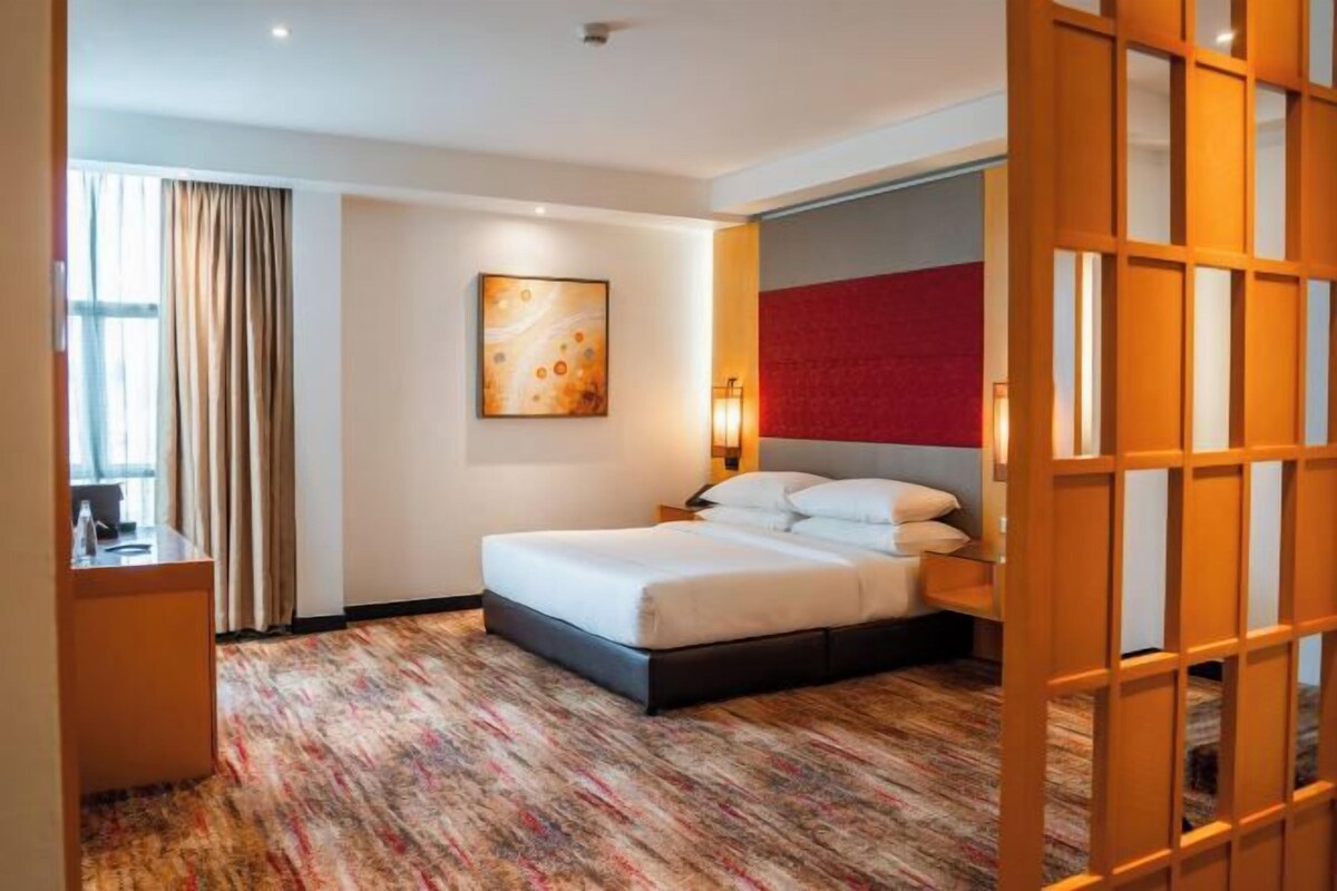 A hotel room with a bed, a desk, a chair, a window with curtains, and a painting on the wall.