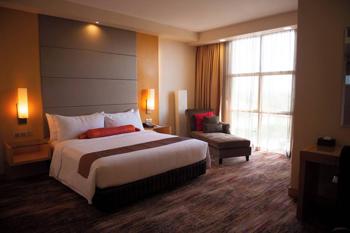 A hotel room with a large bed, a desk, a chair, and a large window with curtains.