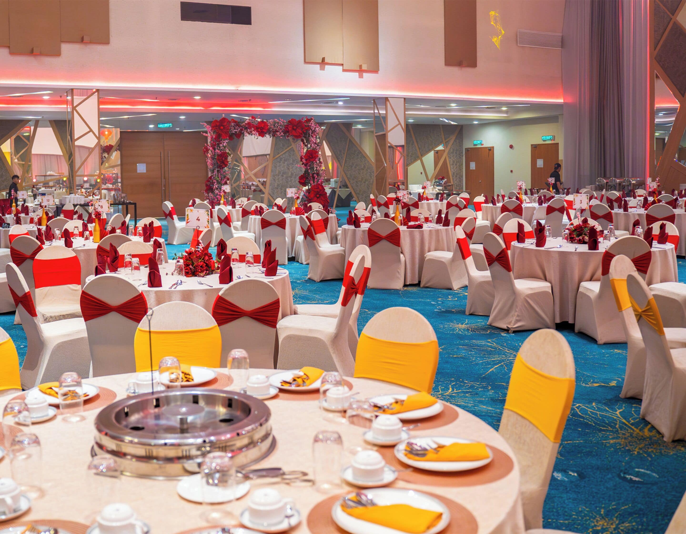 A banquet hall with round tables, white chairs with red sashes, and a blue carpet.