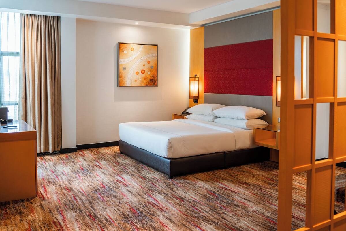 A hotel room with a bed, a desk, a chair, a wall-mounted artwork, a red accent wall, and a patterned carpet.