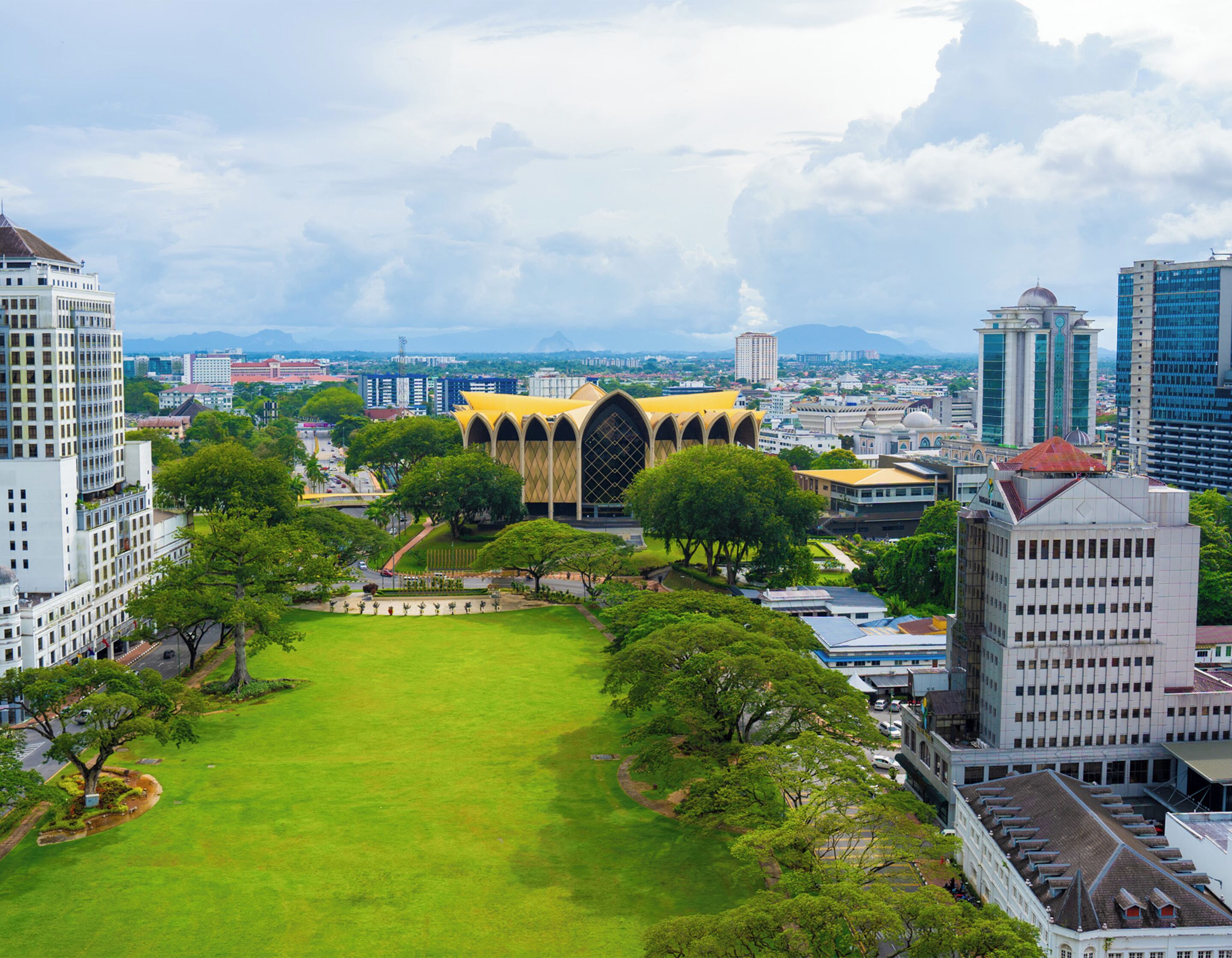 A cityscape with a prominent building featuring a yellow roof and arched windows, surrounded by greenery and other buildings.