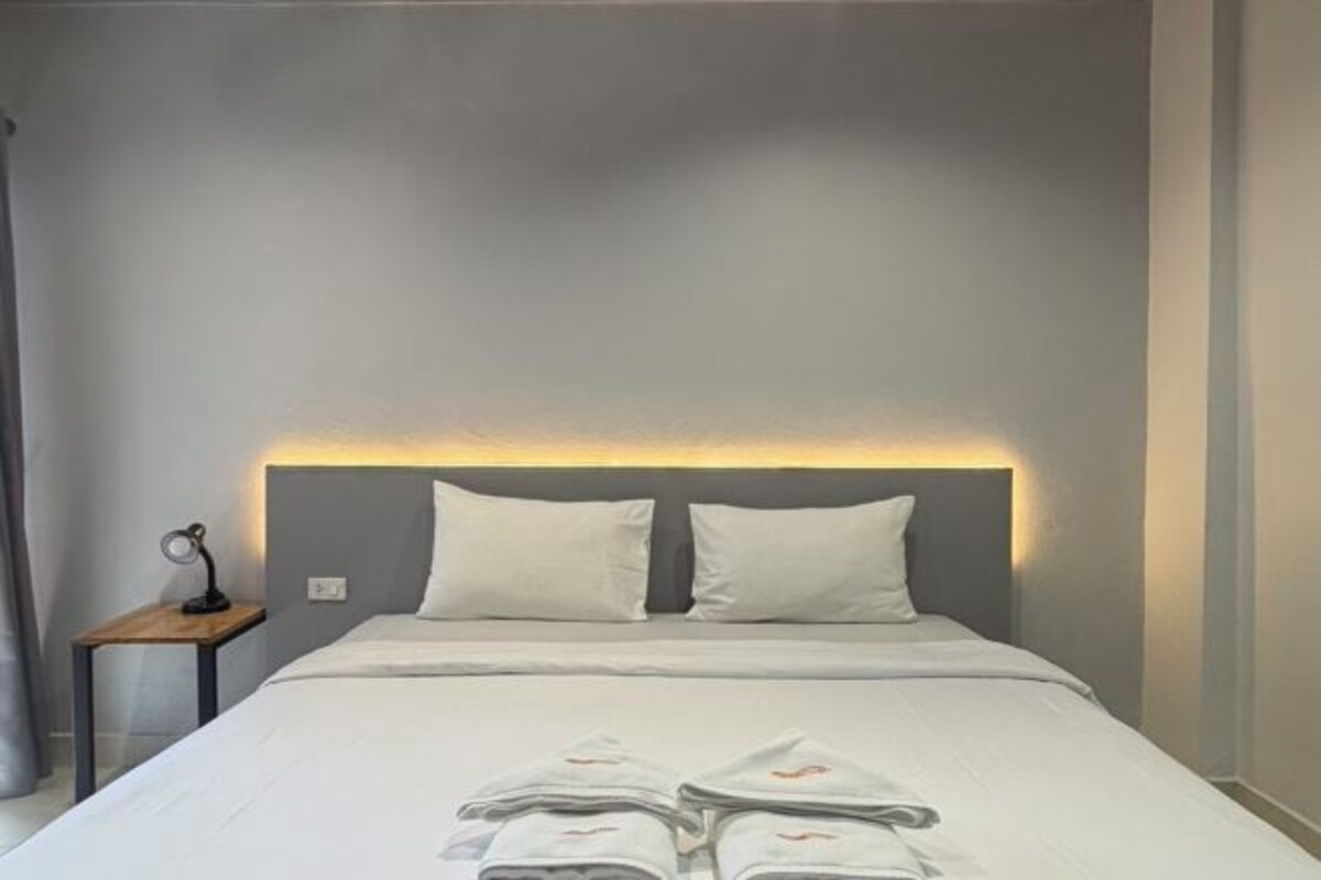 Una cama bien hecha con sábanas y almohadas blancas, una mesita de noche con una lámpara, y un aplique en la pared.