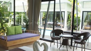 Una habitación de hotel moderna con una cama grande, una zona de estar con una mesa y sillas, y vistas a la piscina y la vegetación exterior.