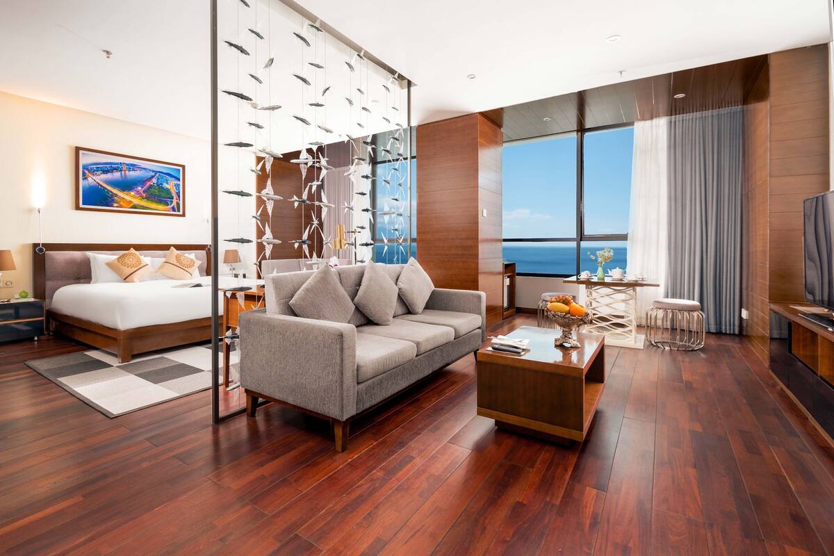 A modern hotel room with a large bed, a sofa, a coffee table, and a view of the ocean.