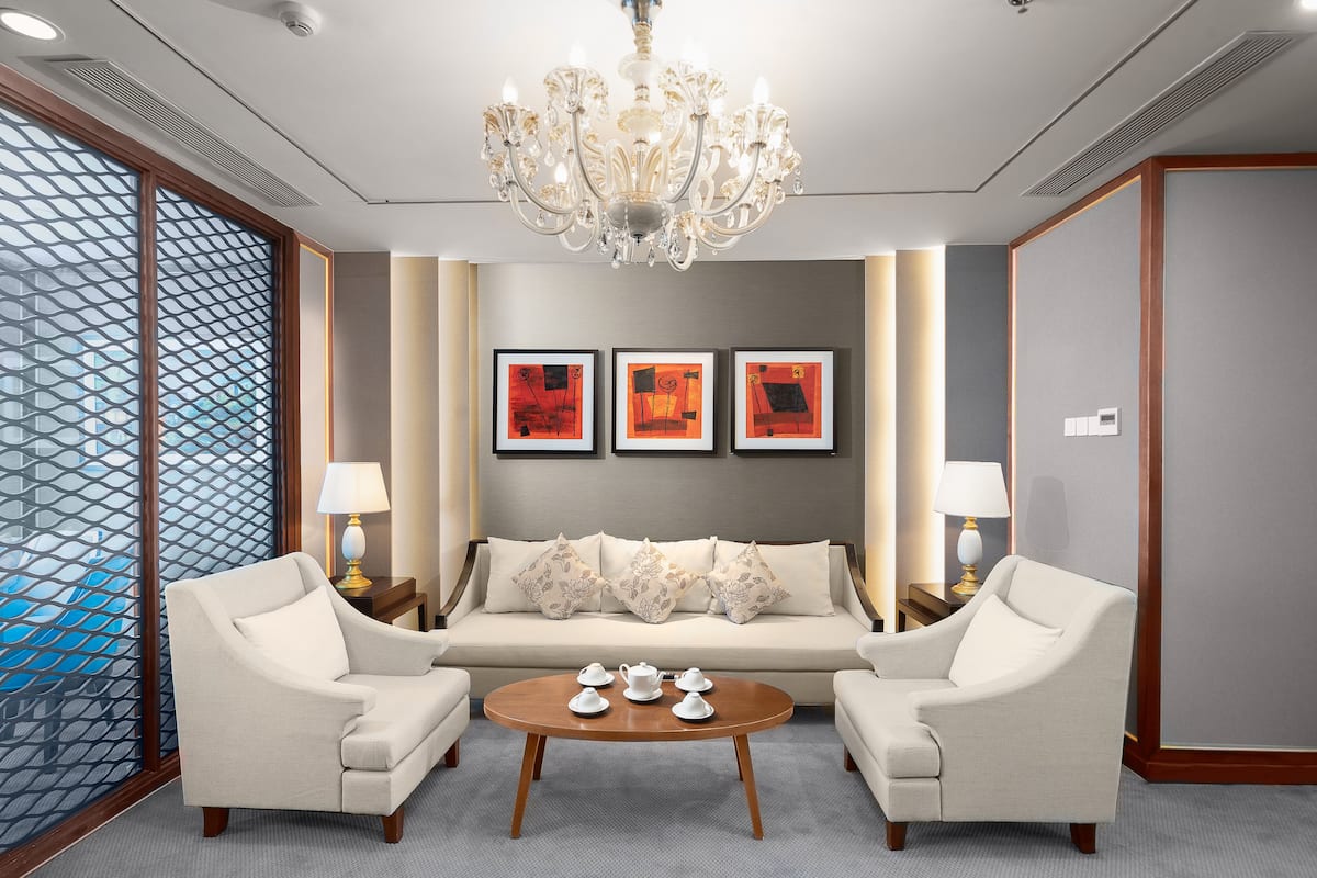 A modern hotel room with a sofa set, two armchairs, a round coffee table, and a chandelier.