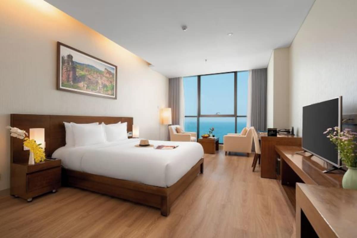 A hotel room with a large bed, a desk, a TV, and a view of the ocean.