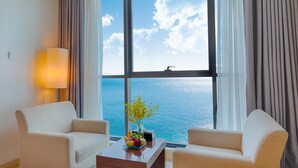 Habitació d'hotel amb dues butaques blanques, una taula de centre de fusta amb fruita i flors, i un gran finestral amb vista al mar.