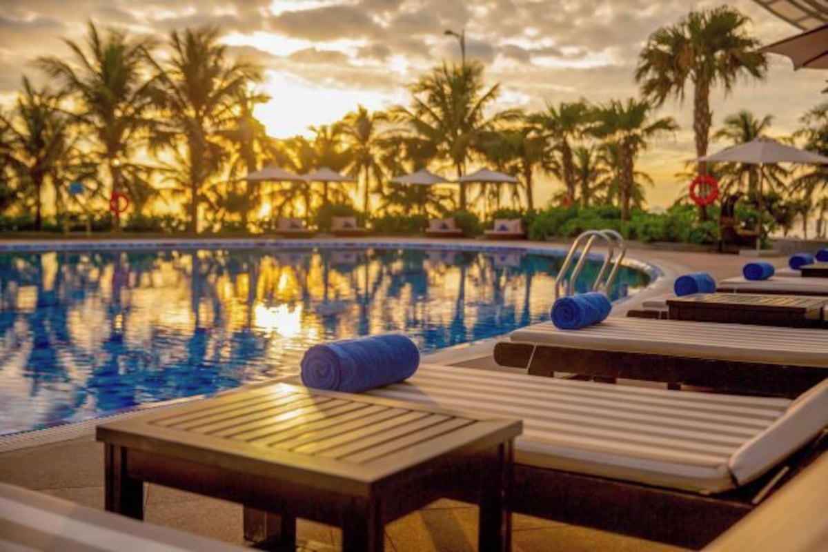 A poolside area with lounge chairs and umbrellas, surrounded by palm trees and a sunset sky.