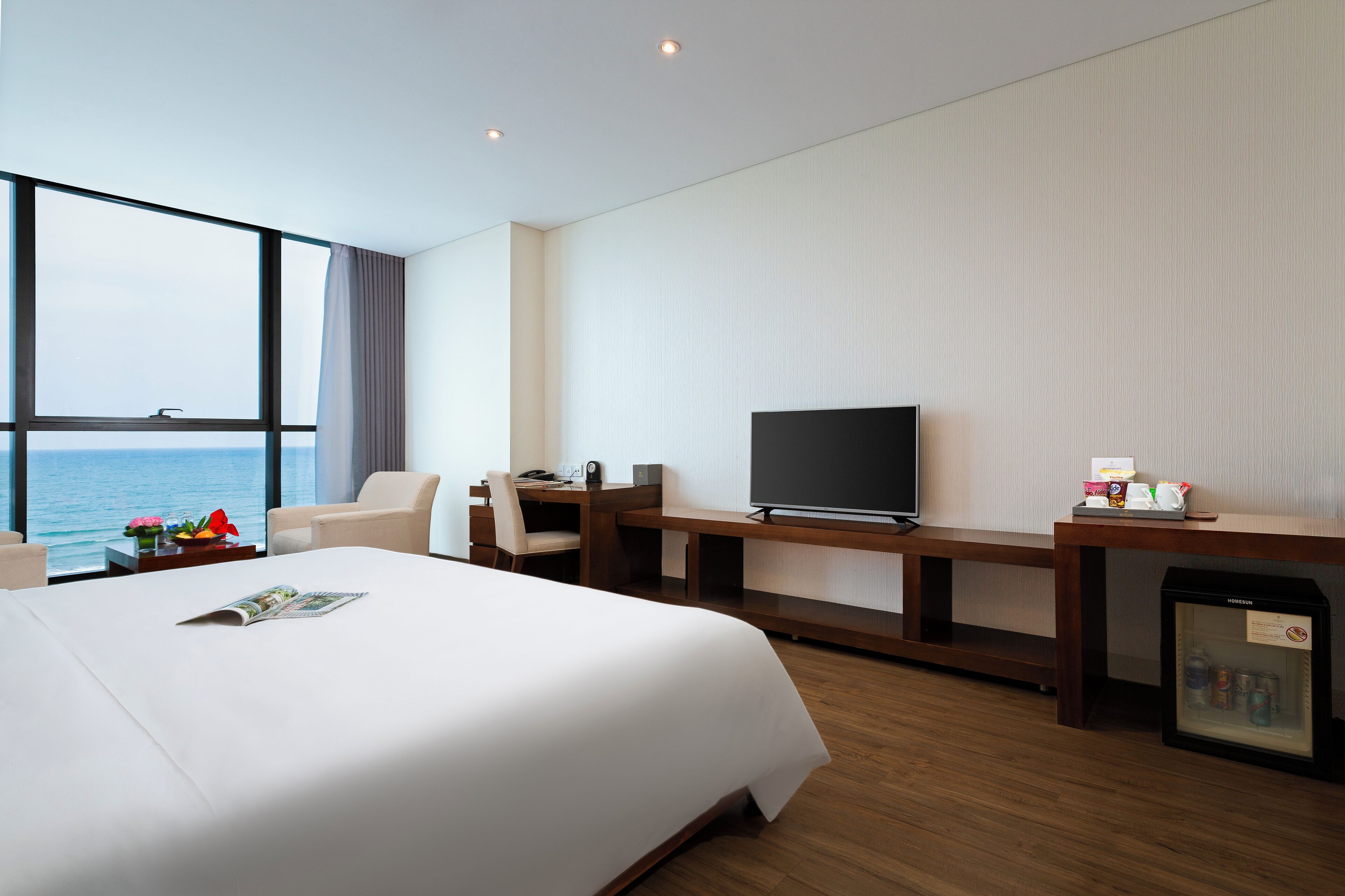 A hotel room with a large bed, a desk with a TV, a small fridge, and a view of the ocean.