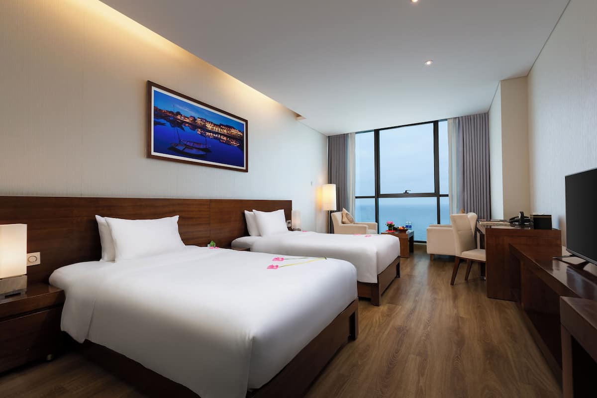 A hotel room with two beds, a desk, a chair, a TV, and a large window with a view of the sea.