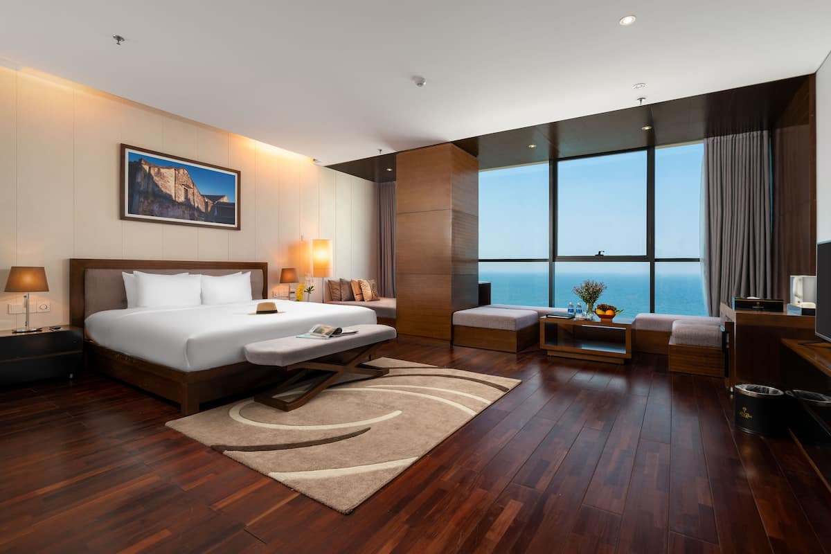 A modern hotel room with a large bed, a sofa, a small table with a vase, and a view of the ocean through large windows.