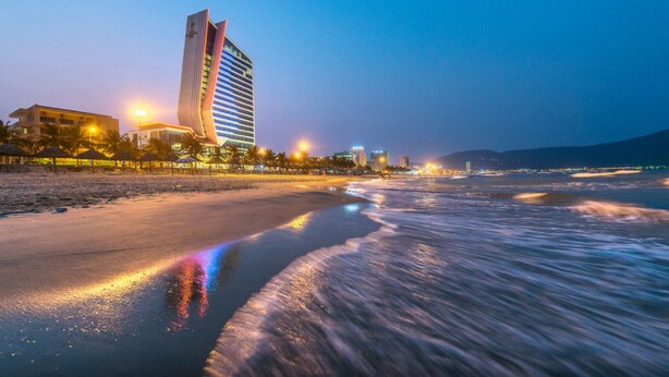 Hotel a peu de platja amb un disseny modern, una platja al primer pla i llums de ciutat al fons.