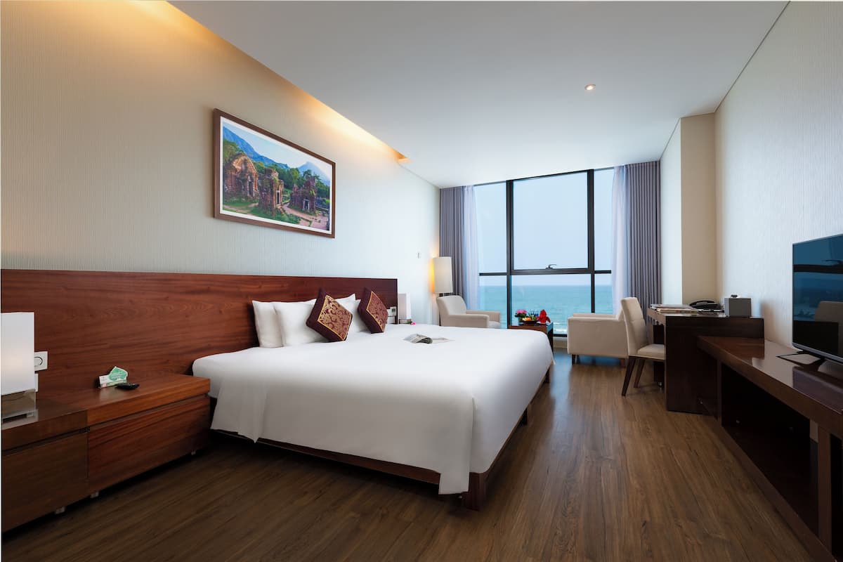 A hotel room with a large bed, a desk, a chair, a TV, and a window with a view of the sea.