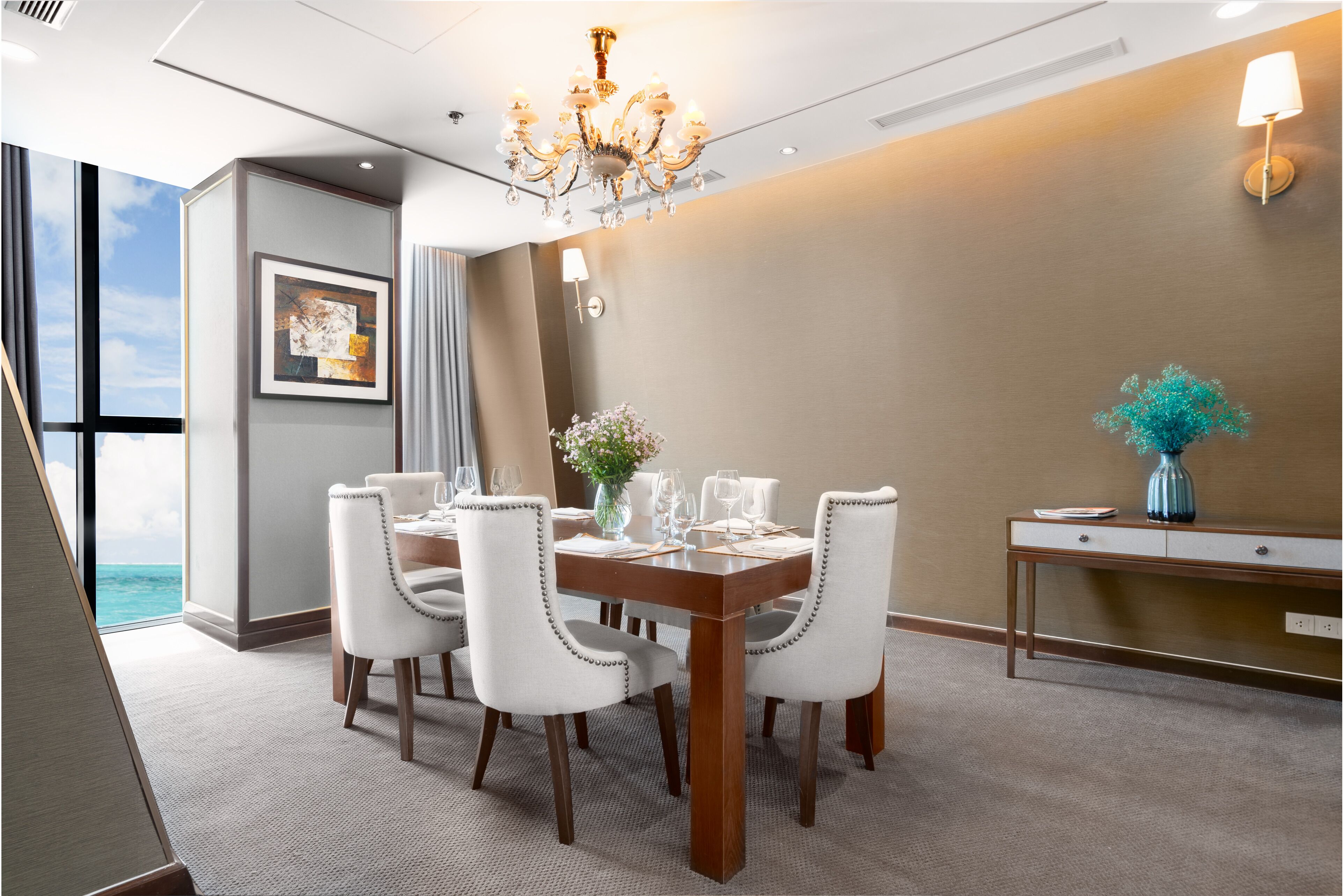 Un comedor con una mesa de madera dispuesta para seis personas, sillas blancas tapizadas, un candelabro y vista al océano.