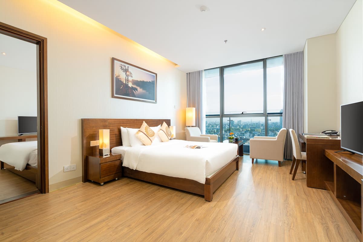 A hotel room with a large bed, a desk, a chair, a TV, and a large window with a city view.