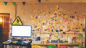 Ein Zimmer mit einer unordentlichen Wand, die mit bunten Haftnotizen, einem Computer und einem Schreibtisch mit diversen Gegenständen bedeckt ist.