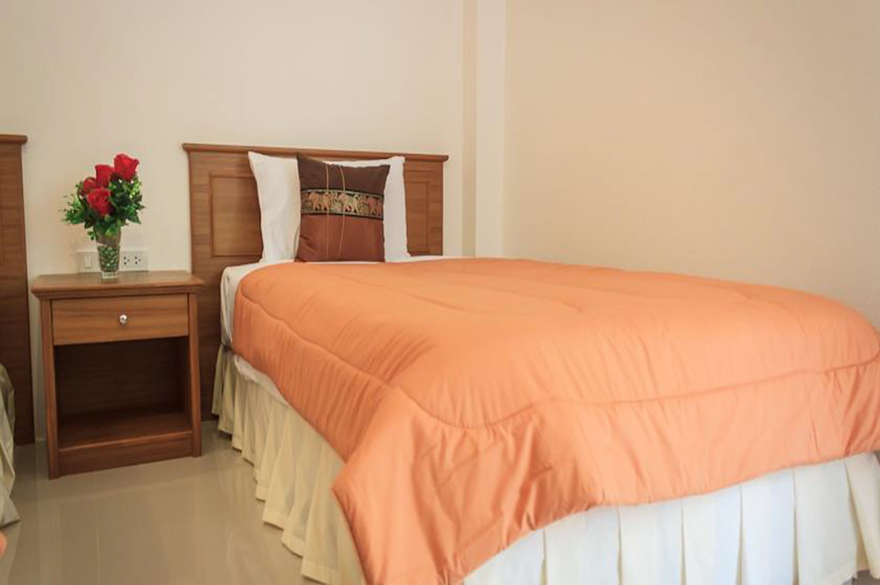 Un letto rifatto con copriletto arancione e cuscini bianchi, un comodino in legno con un vaso di rose rosse, e un arredamento semplice e pulito.