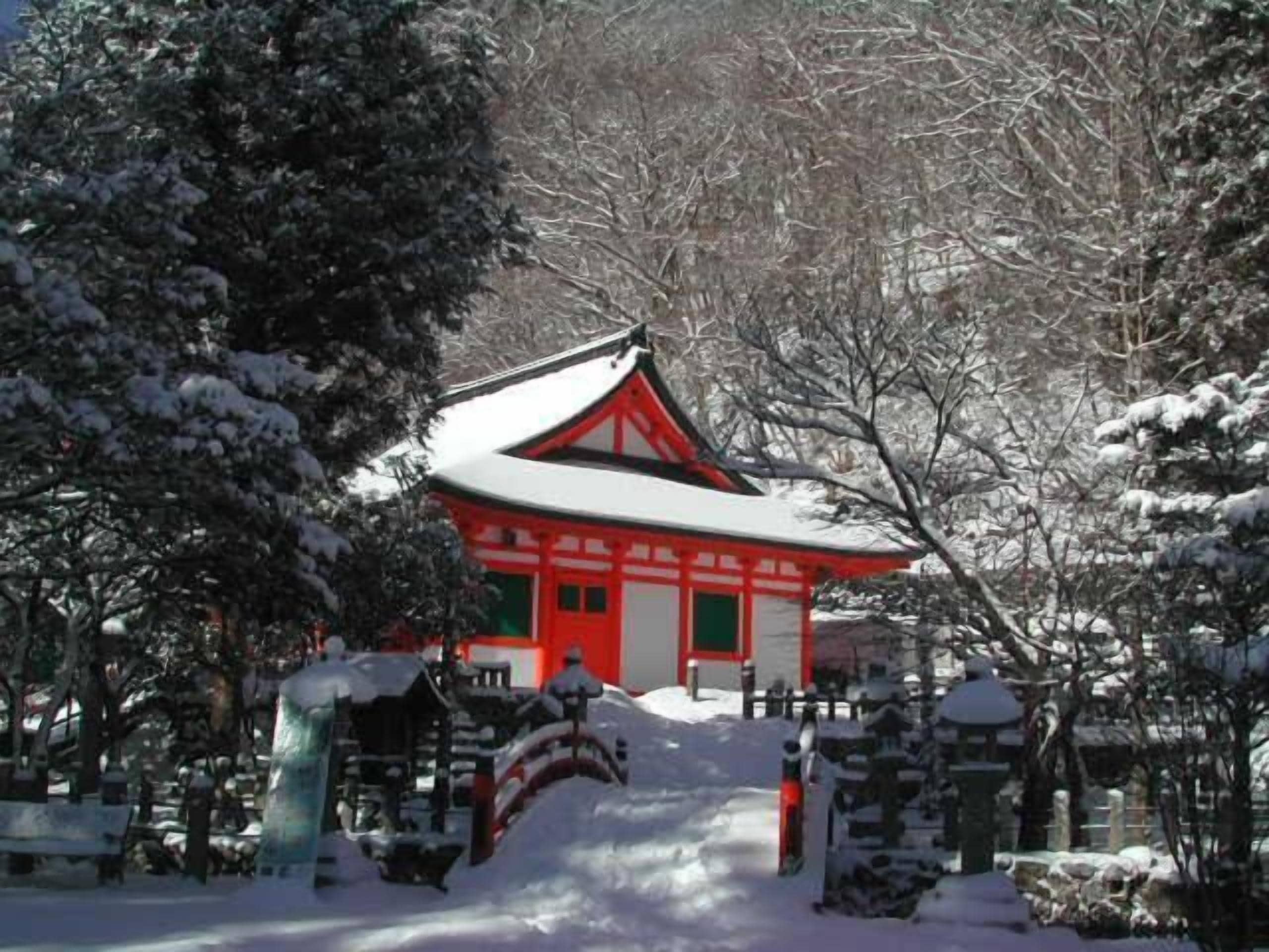 Um edifício japonês tradicional com fachada vermelha e branca, rodeado por árvores cobertas de neve e um pequeno santuário.