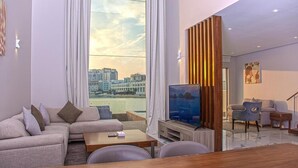 A modern living room with a large TV, comfortable seating, a wooden coffee table, and a view of a cityscape through large windows. -  Sealoft Villas (Amwaj Islands)