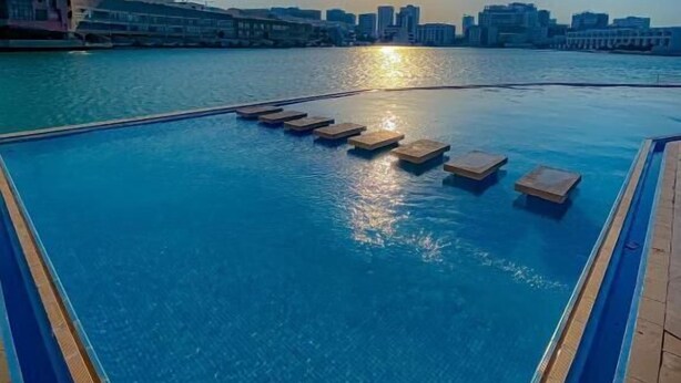 A swimming pool with a view of a city skyline and the sun setting over the water. -  Sealoft Villas (Amwaj Islands)