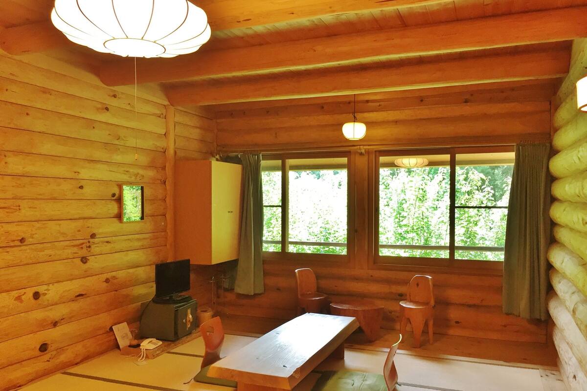 A wooden cabin interior with a dining table, chairs, a television, and a large window with curtains.