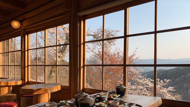 Une table à manger en bois, ornée de différents plats et de théières, se dressant sur fond de fleurs de cerisier et d’un paysage montagneux, le tout visible à travers de grandes fenêtres.